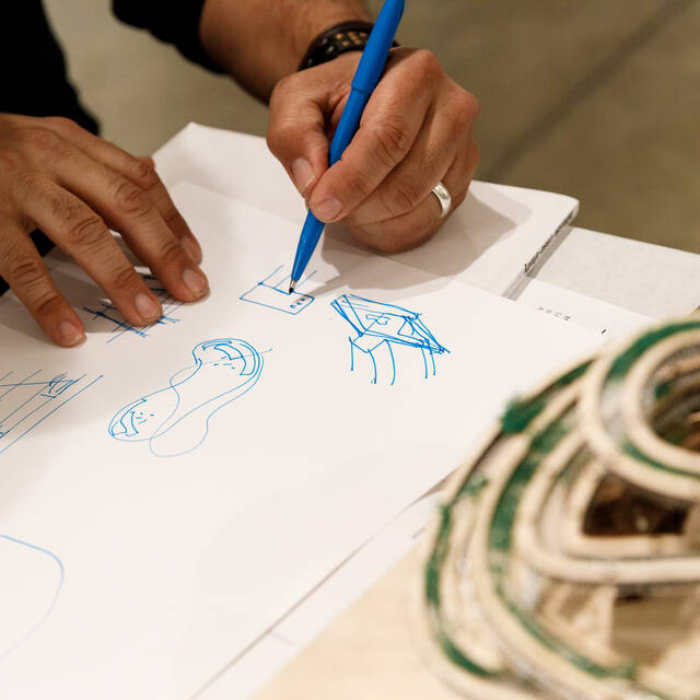 hands drawing on a white sheet of paper with building model nearby