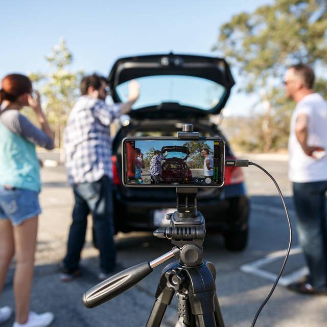 Two men and a woman stand looking into a black hatchback car trunk in front of a Iphone filming them. 