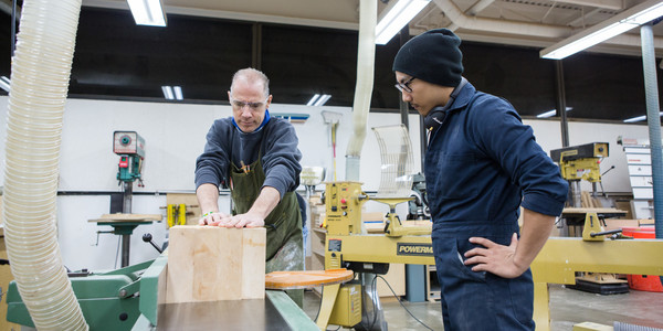 A student looks on as an instructor demonstrates how to cut wood on a a table saw