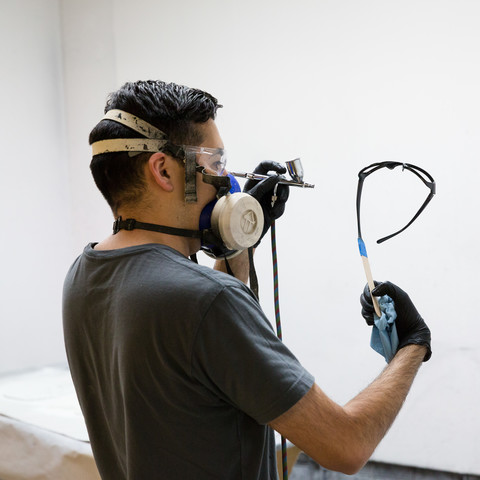 A student wearing a ventilator mask airbrushes eye glass frames. 