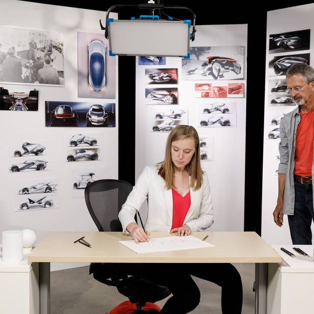 Kayla Rangle drawing in front of transportation design sketches on the wall as a man looks on. 