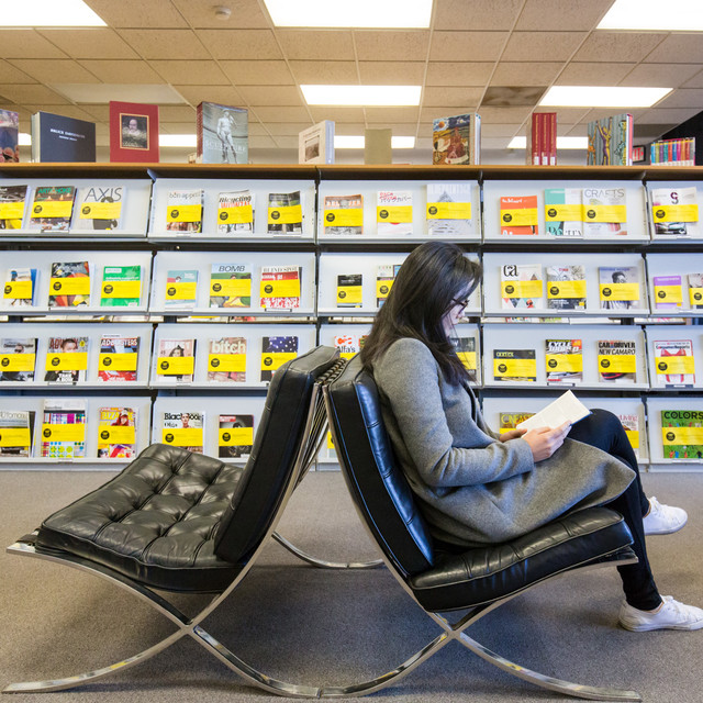An ArtCenter student reads a book while sitting on a black chair in front of the magazine collection