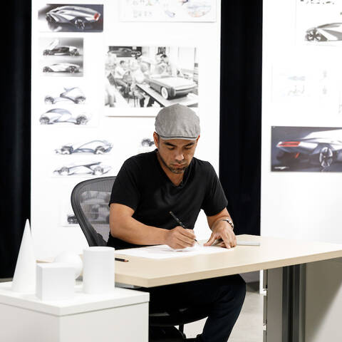 /Instructor Leon Paz drawing at a table with car sketches on a wall behind him