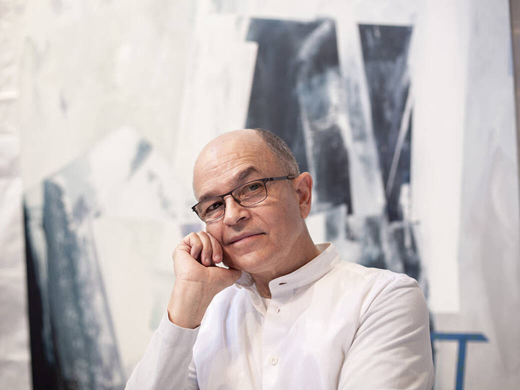 Photograph of Ramone Muñoz, wearing glasses and a long-sleeved white shirt, with a painting in the background.