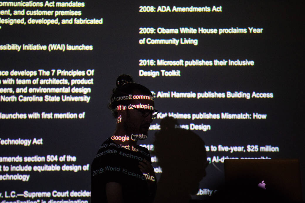 Graphic Design alum, disability rights advocate, designer and Designmatters and Interaction Design Assistant Professor Josh Halstead teaching in a classroom about design accessibility, in front of text from their presentation being projected behind them.