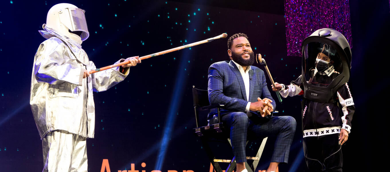 Anthony Anderson, host of the 2021 Make-up Artists and Hair Stylists Guild Awards, on stage at the event and receiving a socially distant make-up touch-up by two individuals in hazmat suits.