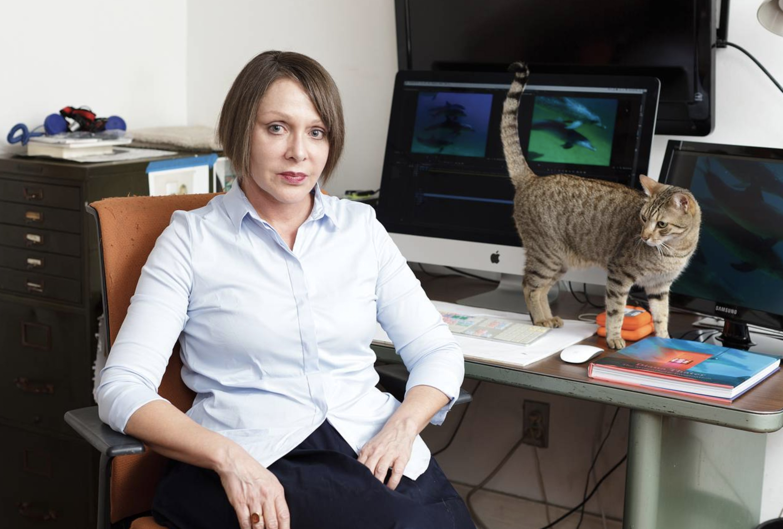 Photo of artist Diana Thater with her cat walking across her work space in front of a computer screen on a desk.