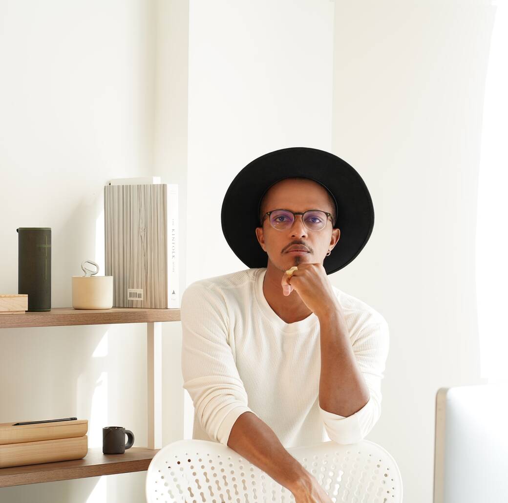 Designer Jeremiah Baker, wearing a white sweater and a black brimmed hat, with mutliple products he