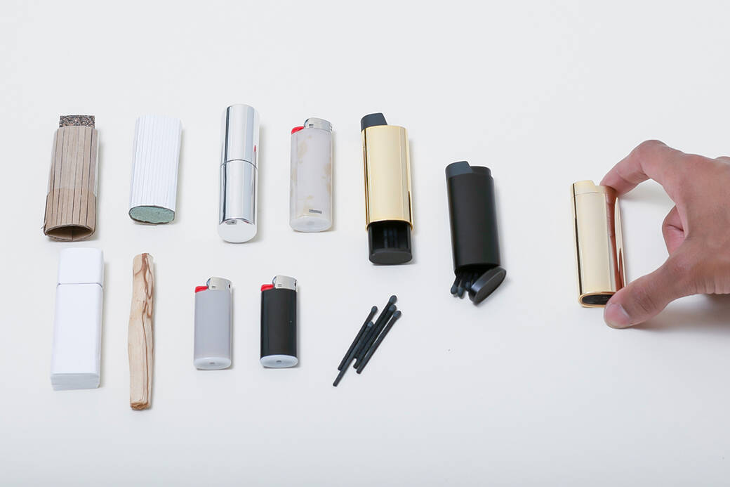 Two fingers hold the Analog Brass Lighter, which lays next to other archetypal objects from which it drew inspiration, including disposable lighters, a lipstick tube and a stick.