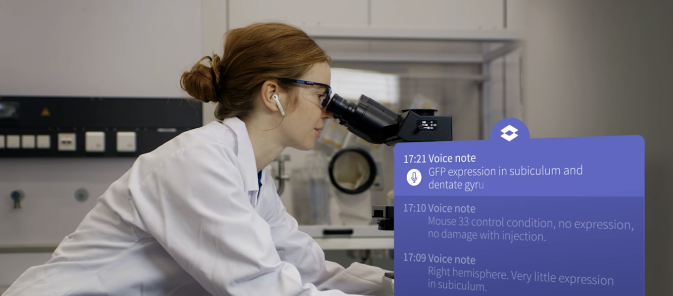 Woman scientist wearing earbuds speaking to the LabTwin digital assistant while looking through a microscope