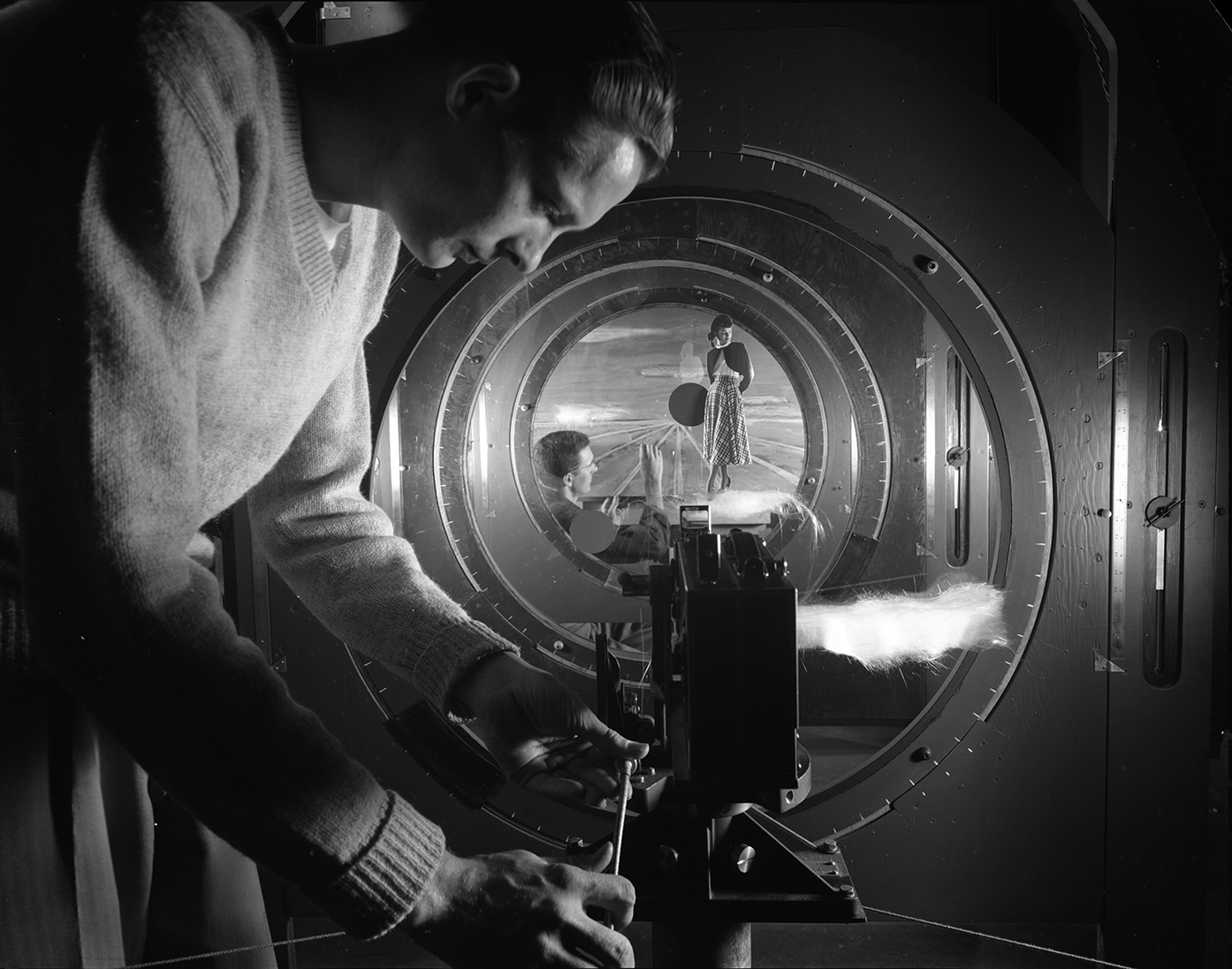 Multilayered black-and-white photograph of two male students working with a multiplane camera, featuring one student in the foreground making camera adjustments, and one student several planes into the image, making adjustments to a painting of a wheat field, over which an image of a woman in a skirt and cardigan has been overlayed