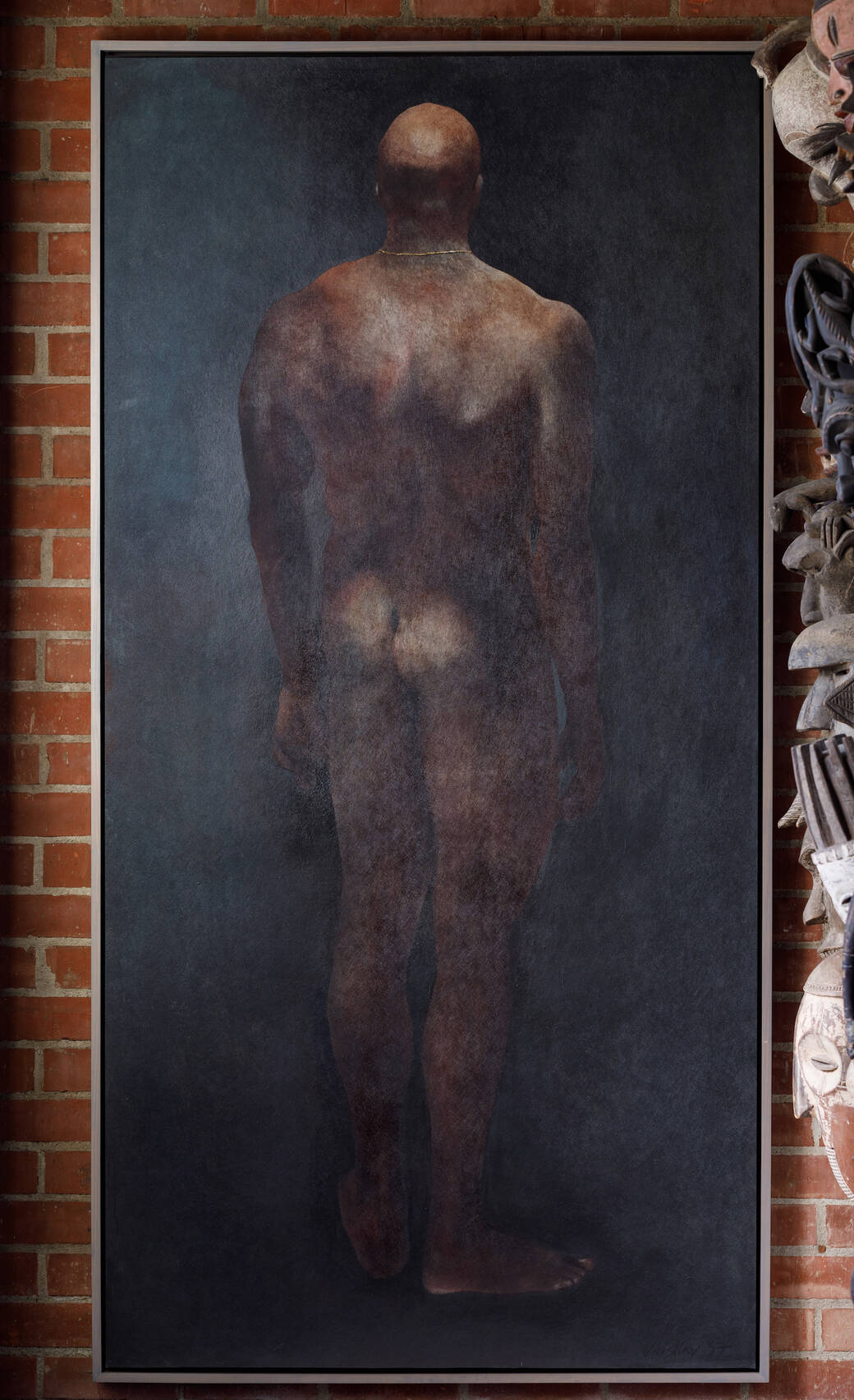 A large painting by Peter Liashkov of a rear view of a nude adult male of African ancestry, photographed against a brick wall and adjacent to masks hanging from a wall, which are partially obscured