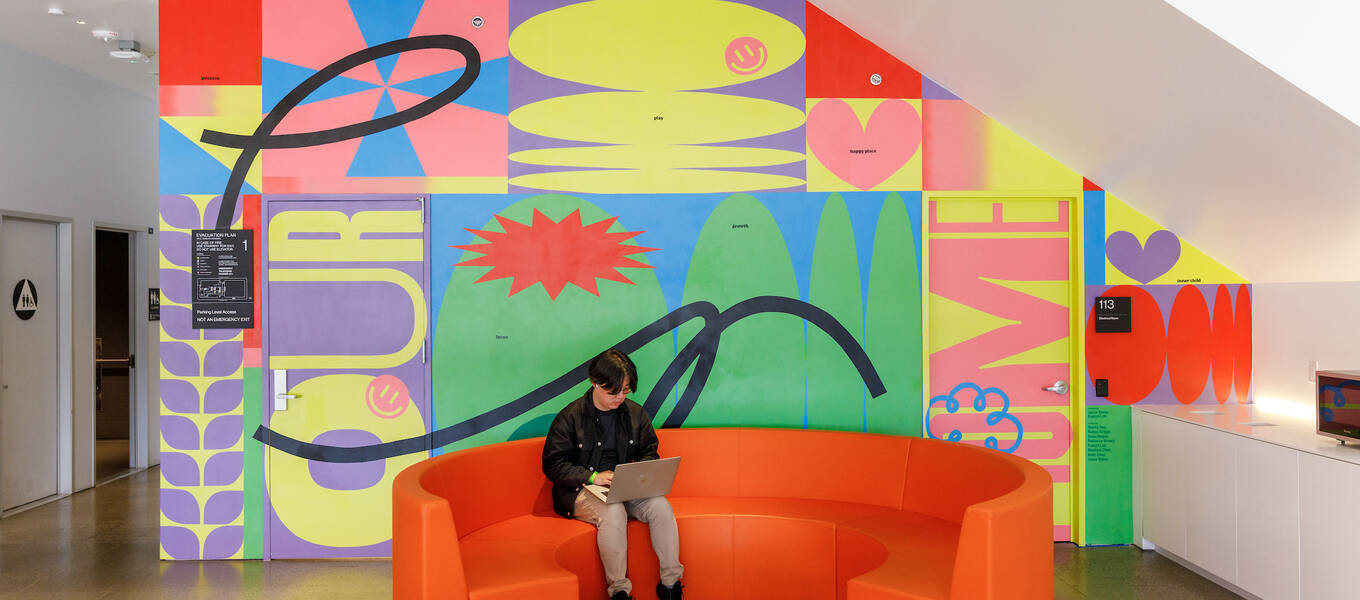 A student sitting in front of a new mural, designed by Graphic Design students Evelyn Luu and Josse Slater, in the lobby of ArtCenter