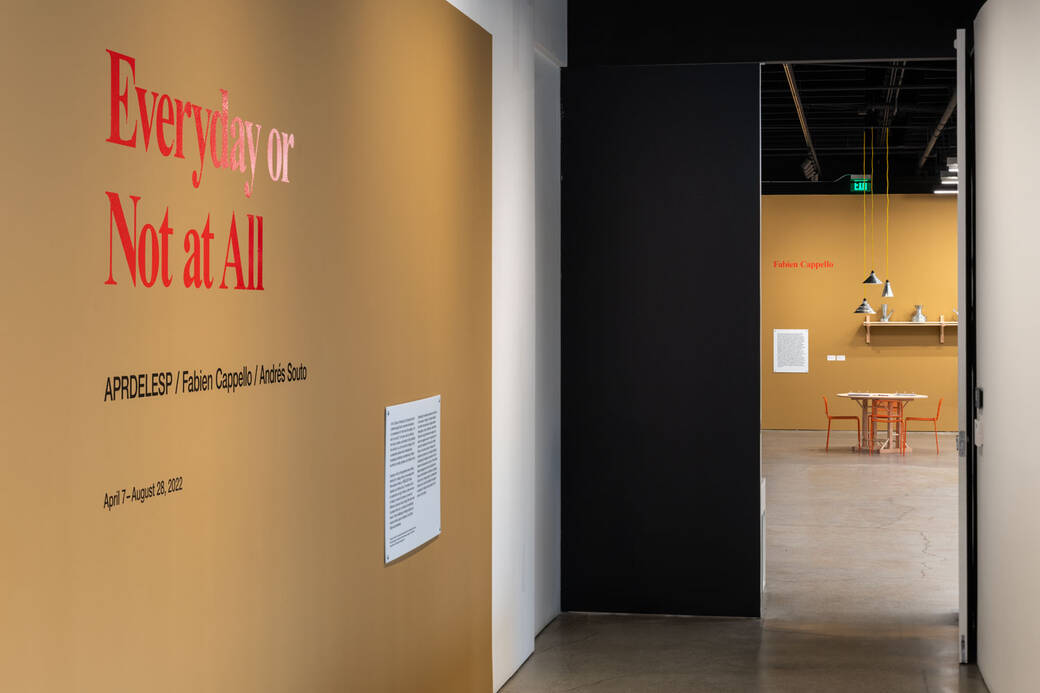 photograph of the entrance to the exhibition of a light brown wall with red letters spelling Everyday or Not at All and the artist names. In the background, you see a table with chairs, silver lights hanging from the exposed ceiling pipes and a shelf on another brown wall.