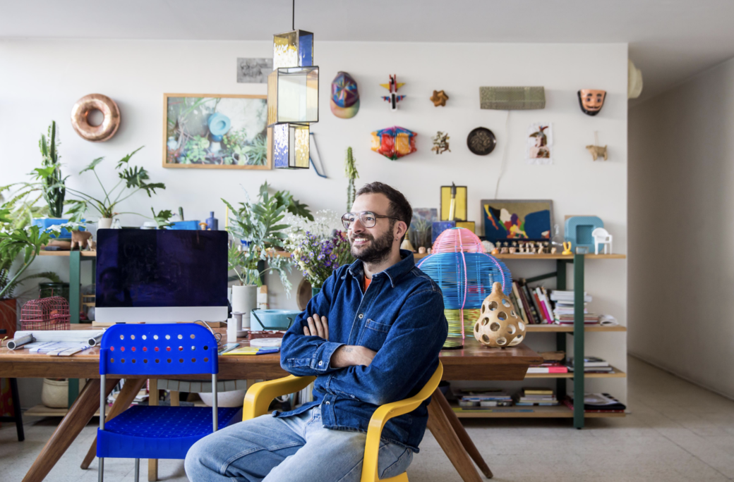 The designer, Fabien Cappello, wears a blue denim shirt and light blue jeans and sits on a yellow chair. In the background is a mac computer on a wooden table with a bright blue desk chair. Beyond that there are many plants, decorations on a wall and a bookshelf. 