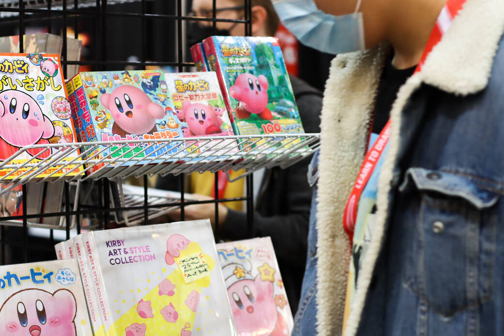 A collection of pink Kirby anime character books on display. There is an attendee wearing a mask looking at them.