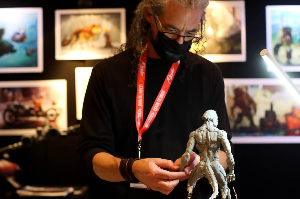 A sculptor giving a live demo at Ligthbox Expo 2022. He is wearing a black mask, sweater, red lanyard and is working on the grey sculpture.