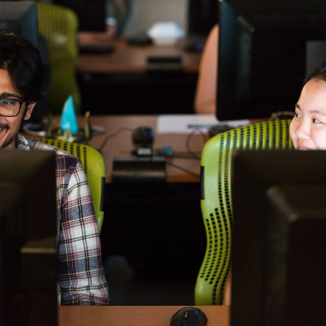 A smiling student in a plaid shirt with glasses smiles at his computer monitor as a neighboring student looks towards him, also smiling. 