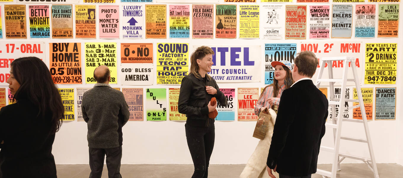 Visitors at the opening of Hello, LA: Clive Piercy, Inside the Mind of a Designer, with a wall covered with posters from the Colby Poster Printing Company and a white stepladder.