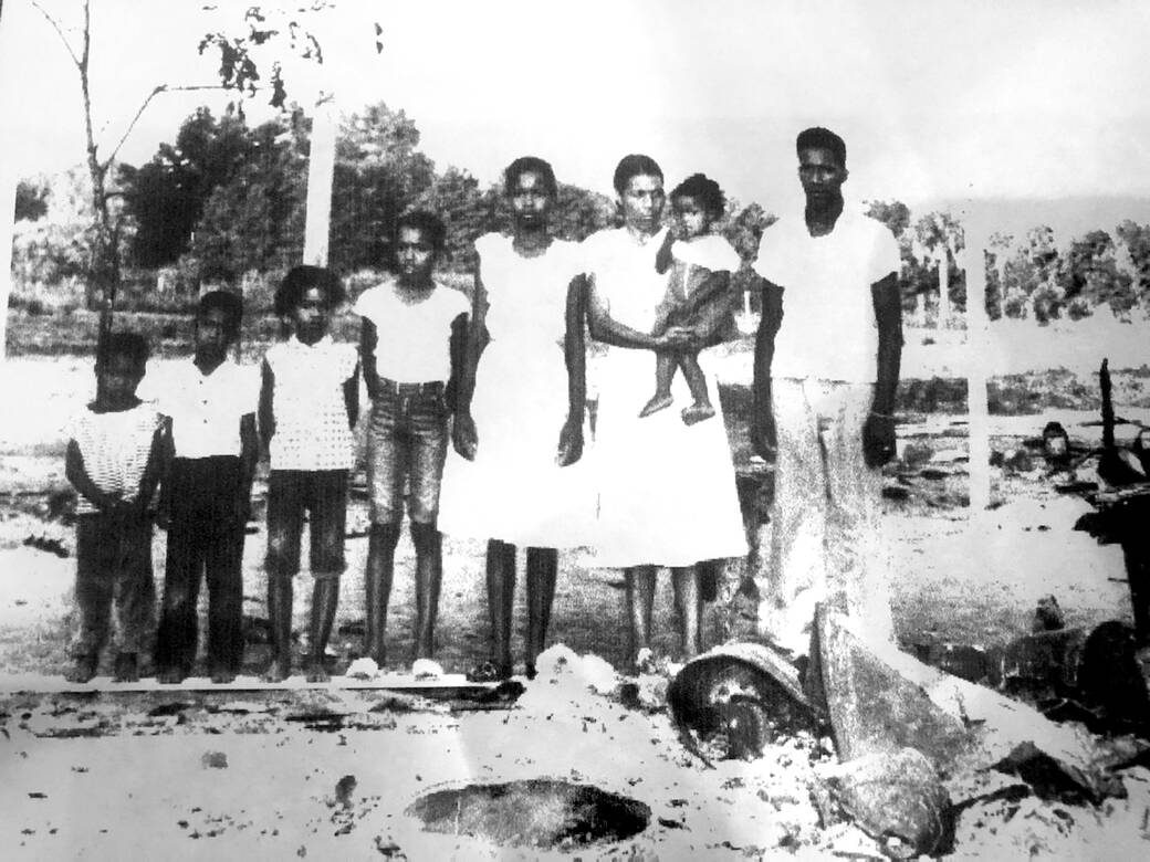 Black-and-white photograph of Kevin Bethune’s family (his mother held by his grandmother) stands in front of the ruins of their home, which was burned down by white supremacists.