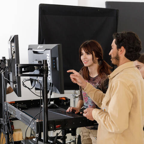 /A student and instructor discuss work on a monitor as another student looks on.