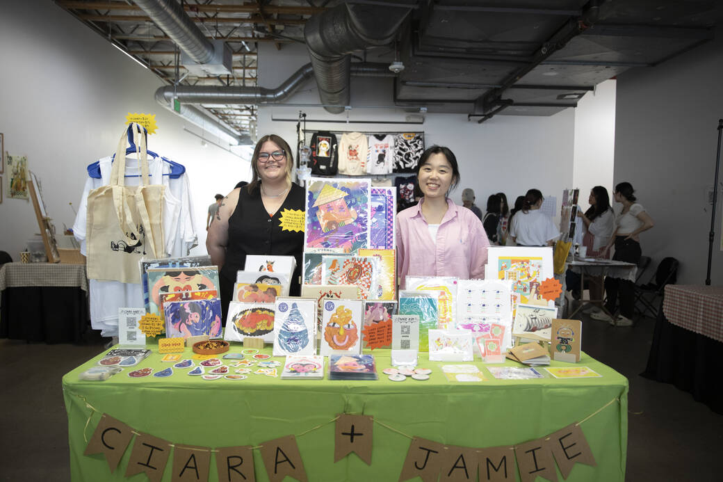 Two individuals selling prints, stickers, buttons and more stand behind a booth labeled Ciara + Jamie at Art Market at Beyond Supersonic