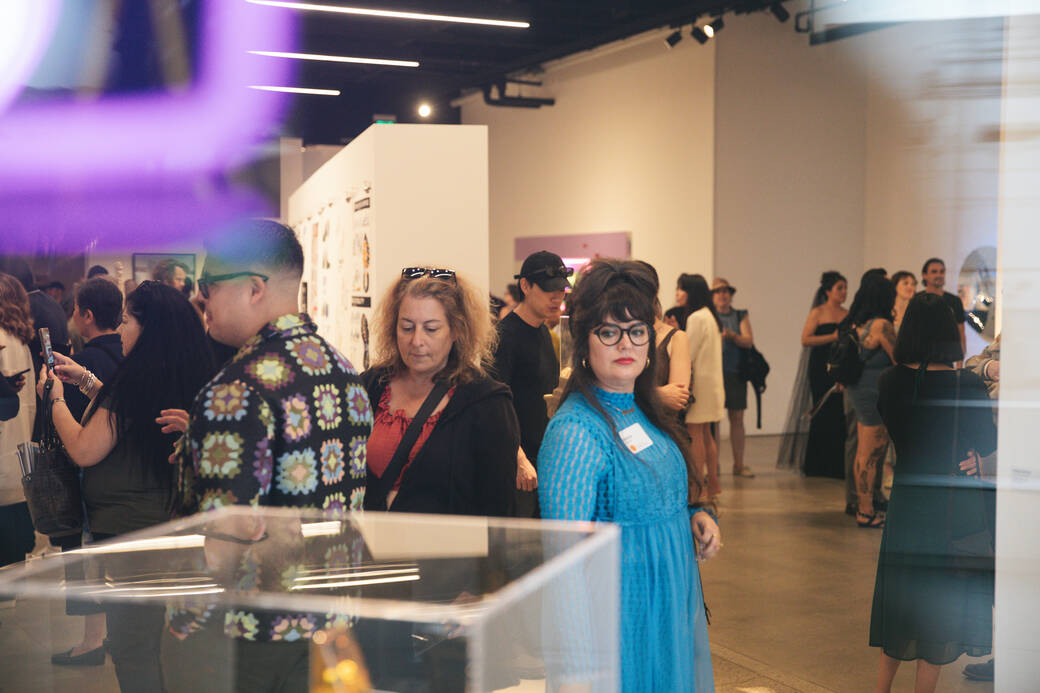 Ramona Rosales, in a blue dress, examines work on display as she walks through a crowded Mullin Gallery at the IdentificarX opening reception.