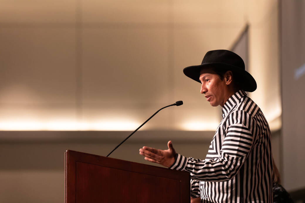 Robert Vargas, wearing a hat and a striped shirt, speaks behind a lectern