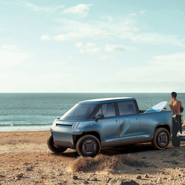 A Telo Truck on the beach, with a surfboard in the truck bed and a shirtless man, looking at the ocean, standing beside it