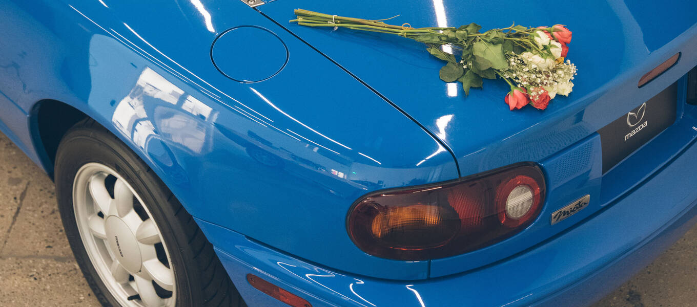 Close-up of a bundle of white and pink roses left atop the closed trunk of a blue 1990 Mazda MX-5 Miata