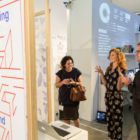 Woman in a black dress gestures excitedly while talking to a man and a woman in front of mfa media design student work