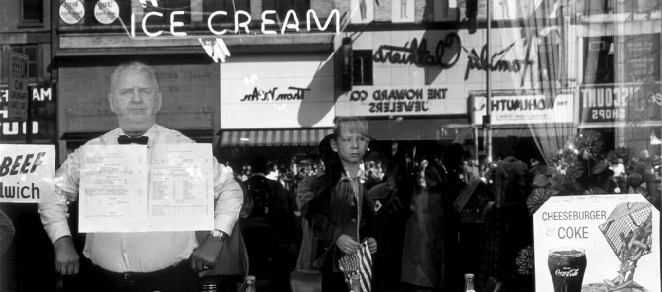 A man and boy inside a diner watch an event, perhaps a parade, unfold outside the restaurant in Lee Friedlander